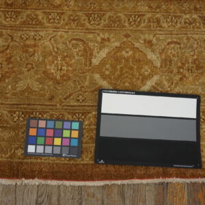 Golden brown high-pile rug featuring an ornate traditional pattern, fringed edges, and color charts on rustic wood flooring.