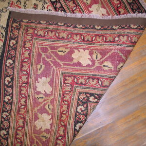 Corner of a low-pile red, beige, and black patterned rug is folded back on hardwood flooring.