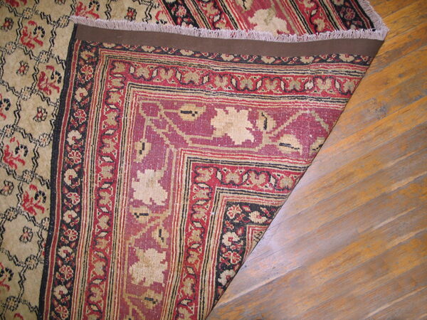 Corner of a low-pile red, beige, and black patterned rug is folded back on hardwood flooring.