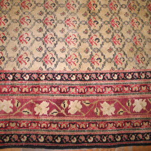 Tan and red patterned rug displaying a repeating field design and detailed borders, visible on wood flooring.