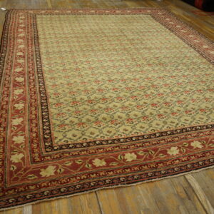 A beige rug with a repeating trellis pattern and a thick red main border is laid on wood planks.