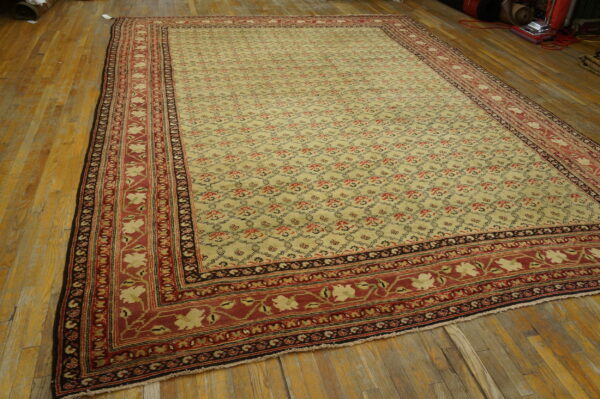 A beige rug with a repeating trellis pattern and a thick red main border is laid on wood planks.