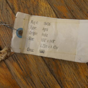 A white product tag is wired to the corner of a dark, patterned rug on warm wood flooring.