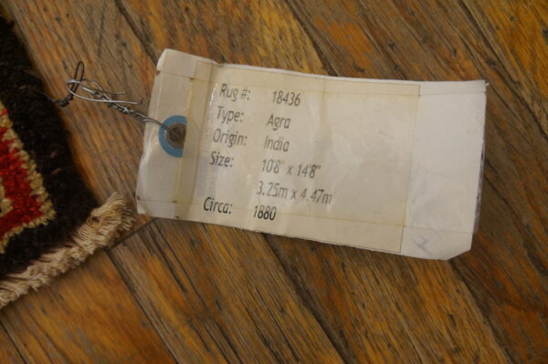 A white product tag is wired to the corner of a dark, patterned rug on warm wood flooring.