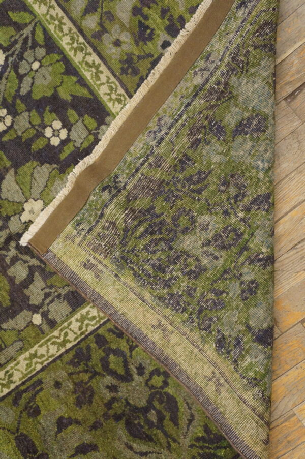 Dark green floral rug folded to show the backing and fringe resting on distressed wood flooring.