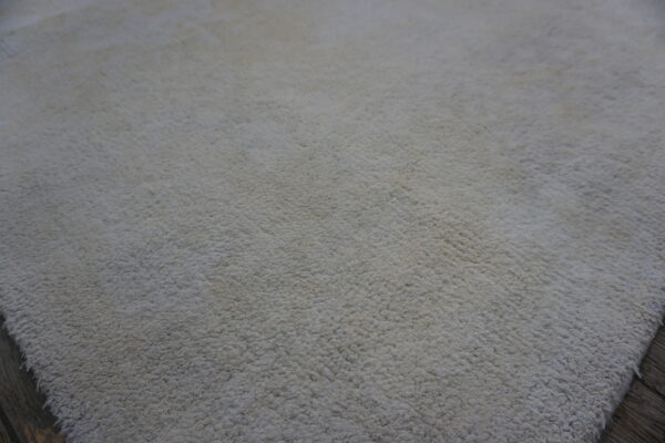 Solid neutral high-pile rug texture closeup, showing edges over dark brown wood floorboards.