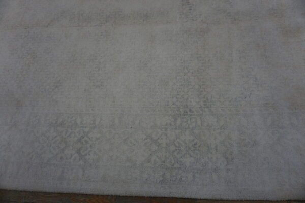 Textured, plush light gray rug with a subtle faded classical design, resting on wood planks.