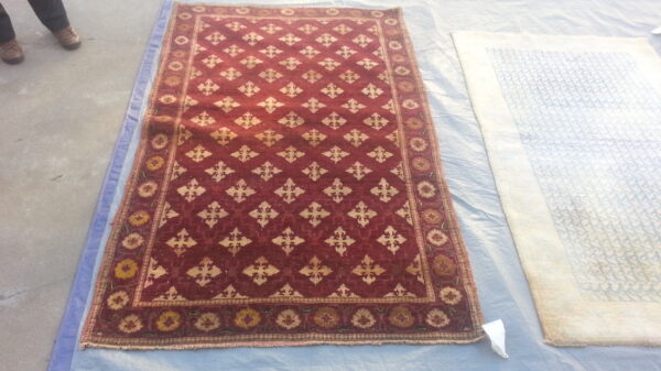 Red patterned area rug laid on a blue tarp next to a lighter rug on a concrete floor.