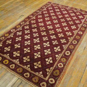 Low-pile burgundy rug with a repeating tan geometric lattice and floral borders rests on worn hardwood floorboards.