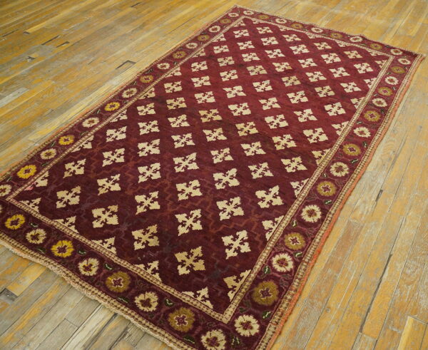 Low-pile burgundy rug with a repeating tan geometric lattice and floral borders rests on worn hardwood floorboards.