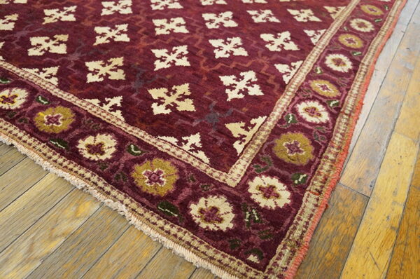 Deep red rug featuring cream geometric motifs and a wide floral border on light wood planks.