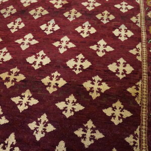 A high-pile burgundy rug featuring cream diamond motifs and a floral border on wood flooring.
