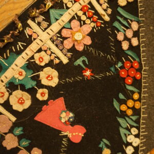 Detailed view of a black folk art rug with appliqué flowers, crosses, and houses next to wood flooring.