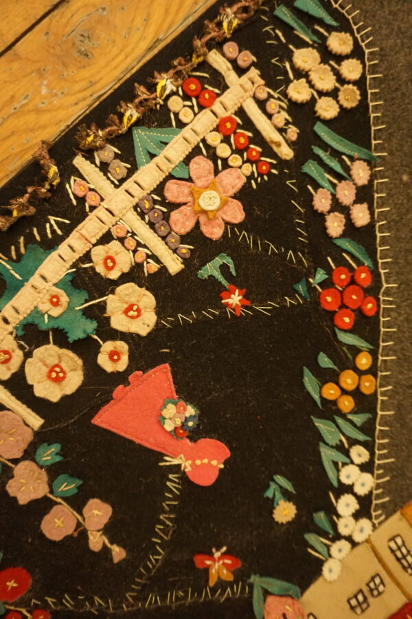 Detailed view of a black folk art rug with appliqué flowers, crosses, and houses next to wood flooring.