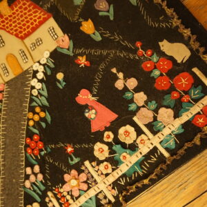 A low-pile black rug featuring colorful stitched appliqué designs of a house, garden, and cat rests on wood flooring.