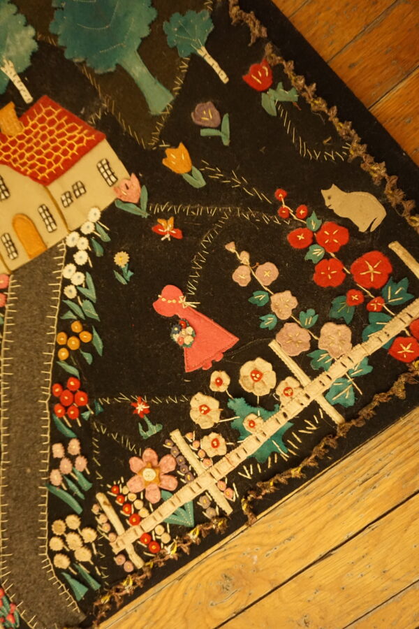 A low-pile black rug featuring colorful stitched appliqué designs of a house, garden, and cat rests on wood flooring.