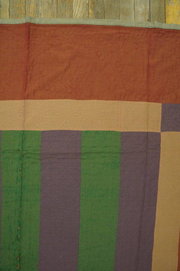 Quilted textile featuring broad stripes of terracotta, tan, green, and mauve laid flat on rustic wood flooring.