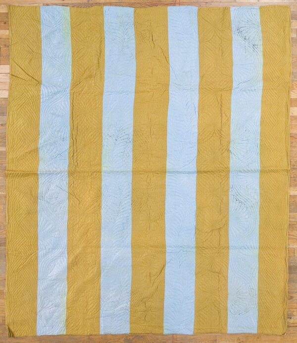 Quilted rug with wide vertical gold and pale blue stripes lies flat on a light wood floor.