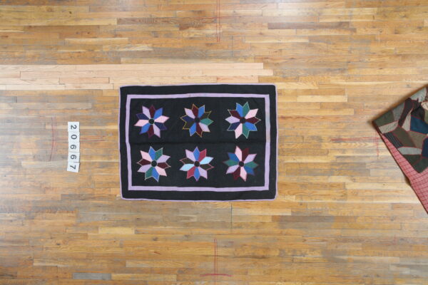 Overhead view of a black rug with multicolored stars and lavender borders, displayed on light wooden floorboards.