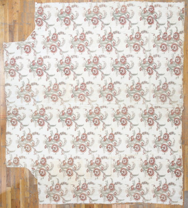 Creamy textile with repeating floral motifs in rust and sage green, laid on hardwood flooring.