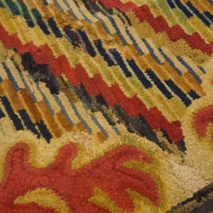 Textured rug close-up with thick red, gold, navy, and green abstract patterns over wood planks.