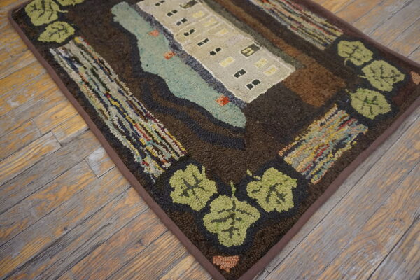 A pictorial hooked rug featuring a beige building, blue water, and green leaf border on distressed wood floors.
