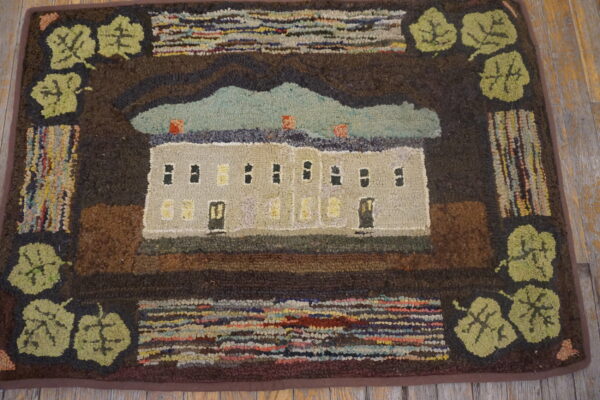 Primitive hooked rug featuring a central house and sky scene bordered by dark brown and green leaves on wooden flooring.