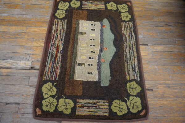 Hooked rug with a beige building, teal water, and olive leaf border on distressed wood plank flooring.