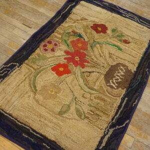 Beige, high-texture hooked rug with red and green florals and a dark blue border rests on worn wood floors.