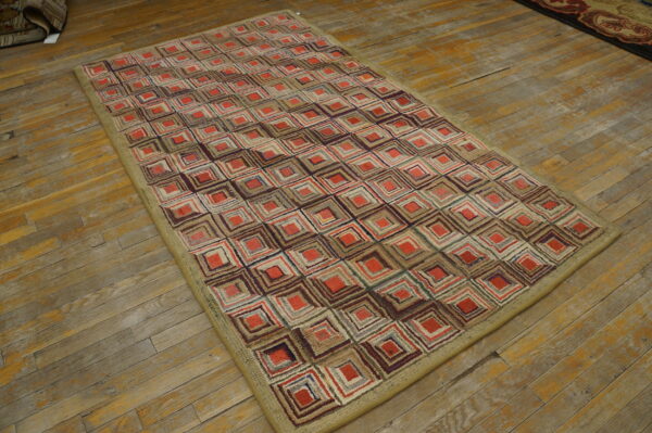 Low pile rug with red, brown, and beige concentric square patterns and a wide khaki border on wood floorboards.