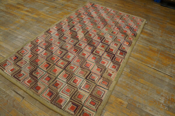 Textured low-pile rug with concentric red and multicolored squares and a khaki border on worn wood floors.