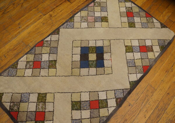 Low-pile rug with a quilt block design in tan, blue, red, and green rests on a hardwood floor.