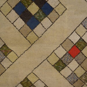 Thick beige rug with a diamond pattern filled by multicolored squares, resting next to wood flooring.