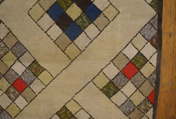 Thick beige rug with a diamond pattern filled by multicolored squares, resting next to wood flooring.