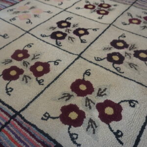 Looped cream rug with a black grid pattern, featuring maroon and pink flowers and a colorful striped border.