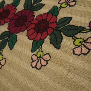 Close view of a nubby tan rug with large red, pink, and green outlined floral designs.