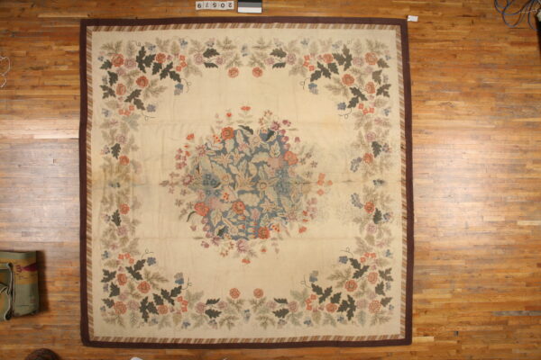 Square rug with a cream floral field, blue-gray central bouquet, and wide dark brown border on wood planks.