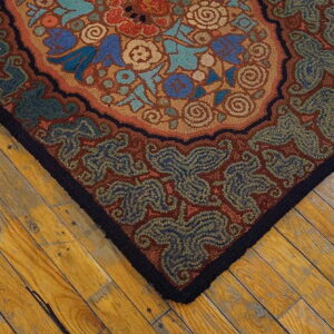 Rust, blue, and teal floral hooked rug corner with a black border on warm wooden plank flooring.