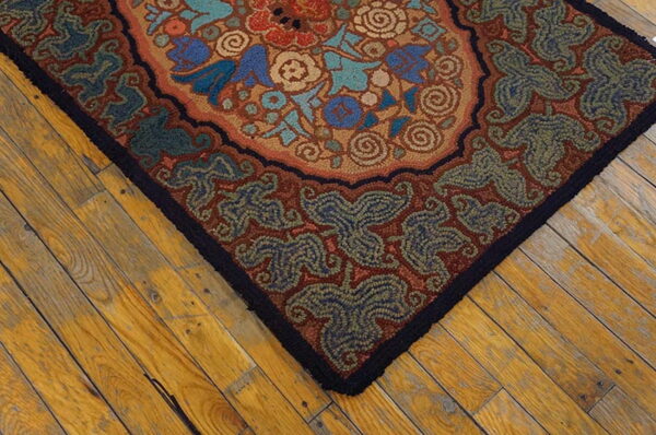 Rust, blue, and teal floral hooked rug corner with a black border on warm wooden plank flooring.