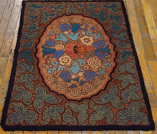 A coarse, hooked-style rug displaying an oval floral pattern in blue, red, and tan on rustic wood floors.