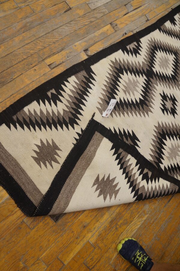 Geometric off-white, brown, and black flatweave rug on worn wood planks, next to a person's foot.
