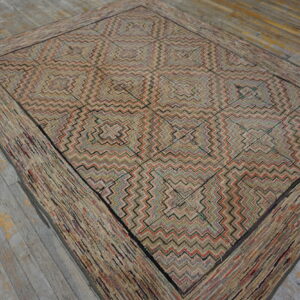 A low-pile rug with concentric zigzag squares and a wide rag-style border rests on distressed wood flooring.