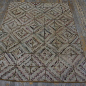 A textured rug with multicolored zig-zag diamonds on a light background rests on worn gray wood flooring.