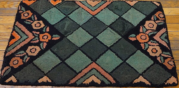 A hooked rug featuring dark green diamonds and orange floral motifs on a black border over wood flooring.