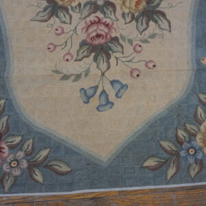 Floral rug with a beige field, blue border, pink and yellow roses, lying on exposed rustic wood floors.