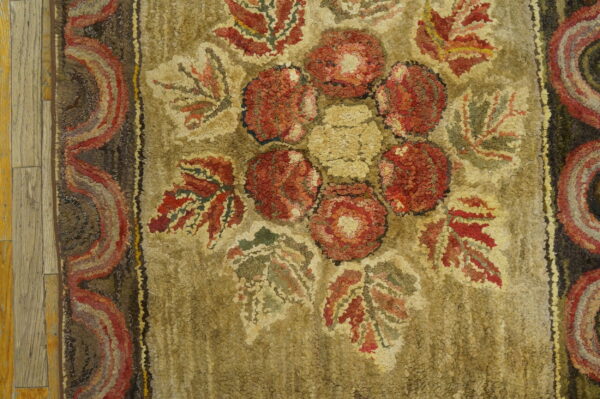 Khaki hooked rug with red floral medallion and striped border detail on light wood planks.