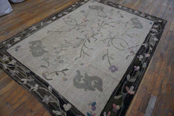 A cream hooked rug featuring sparse floral vines and gray scrolls, framed by a dark, floral border on wood floors.