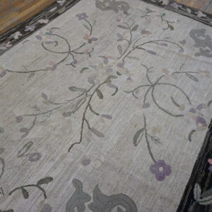 Beige hooked rug with pale purple and gray floral vines, framed by a thick dark patterned border on wood floor.