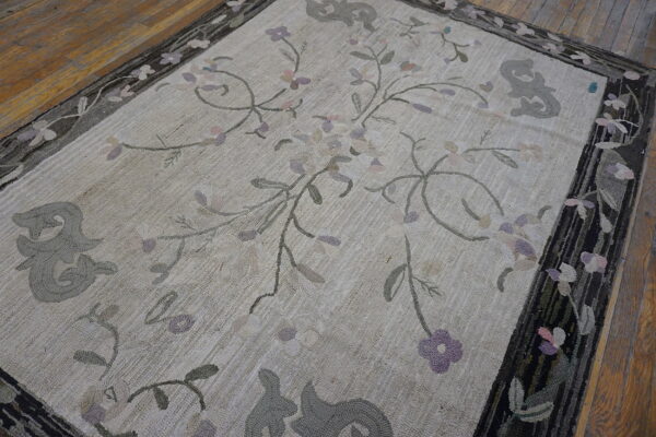 Beige hooked rug with pale purple and gray floral vines, framed by a thick dark patterned border on wood floor.
