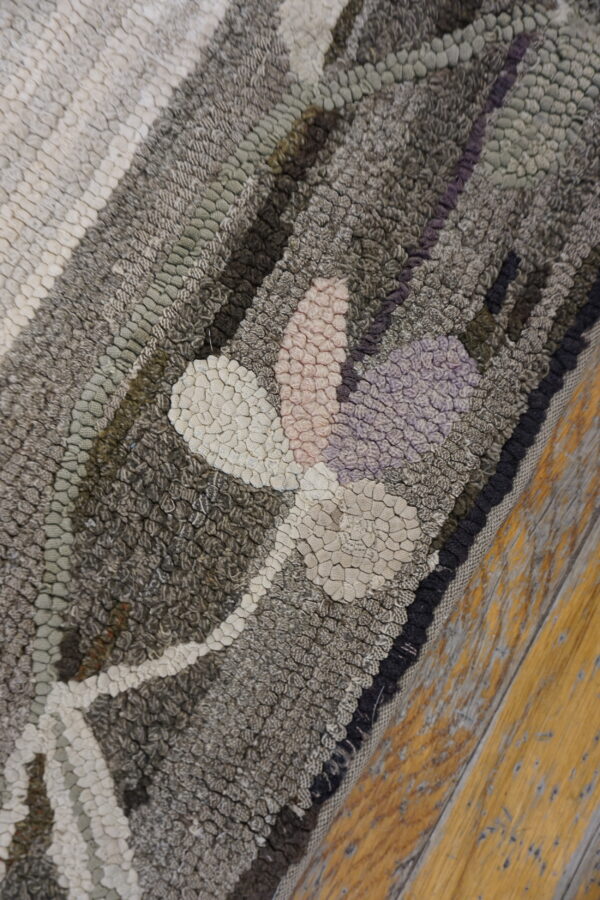 Textured hooked rug detail featuring gray, cream, pink, and purple floral motifs on distressed wood flooring.